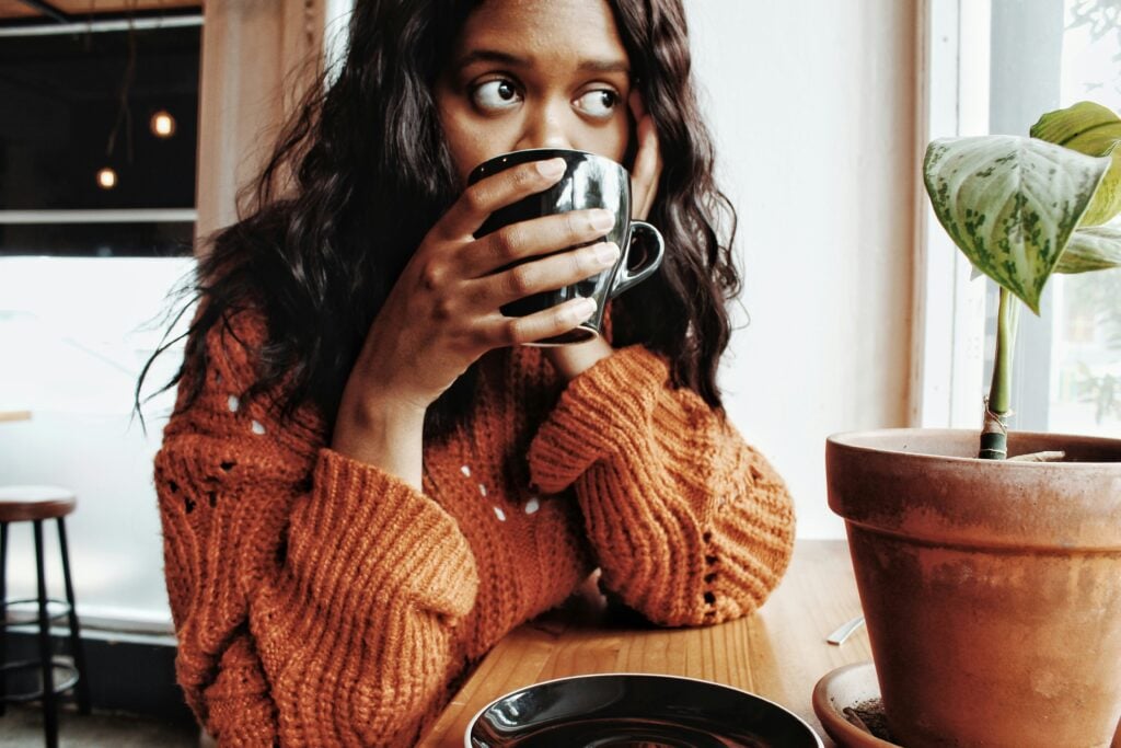 Black woman sitting at a café table holding a mug, looking sideways — self-care for women without money doesn't require a spa day.